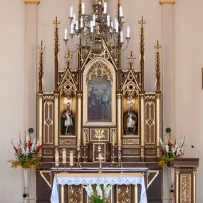 Zdjęcie nr 1: Ołtarz przyścienny, architektoniczny, pięcioosiowy, jednokondygnacyjny ze zwieńczeniem. Antepedium dekorowane płyciną w profilowanej ramie z umieszczonym centralnie czteroliściem oraz krzyżem. Tabernakulum metalowe, jednodrzwiczkowe z umieszczonym centralnie kielichem z hostią na tle kłosów oraz winorośli. Po bokach umieszczone litery „A” i „Ω” na tle krzyża każda. Nastawa posadowiona na wysokim cokole z płycinami wypełnionymi maswerkiem. Osie w układzie schodkowym z osią środkową wyższą i szerszą od bocznych, wydzielone wiązkami służek na postumentach w formie skręconych trzonów, z kapitelami dekorowanymi motywem ostroliścia. Służki zwieńczone pinaklami zakończonymi kwiatonami, w osi środkowej ażurowa wieżyczka w formie zbliżonej do pinakla zakończonego kwiatonem i krzyżem, tworząca baldachim nad figurą Dzieciątka Jezus. Osie środkowe zwieńczone trójkątnymi wimpergami wypełnionymi dekoracją maswerkową. W polu ołtarza obraz Trójcy Świętej w złoconej, profilowanej ramie w kształcie stojącego prostokąta zamkniętego łukiem ostrym z maswerkiem. Poniżej płycina z płaskorzeźbionym krzyżem na tle kłosów. W osiach drugiej i czwartej złocone nisze mieszczące figury św. Stanisława i Wojciecha. Nisze zamknięte od góry muszlami palmetowymi. Pod niszami płyciny wypełnione maswerkiem z fantazyjnym motywem geometrycznym. Osie skrajne wypełnione zdwojonymi blendami z rozetami pośrodku. Struktura ołtarza malowana na brązowo z elementami czerwieni, detal architektoniczny złocony. Figury w pełni polichromowane.