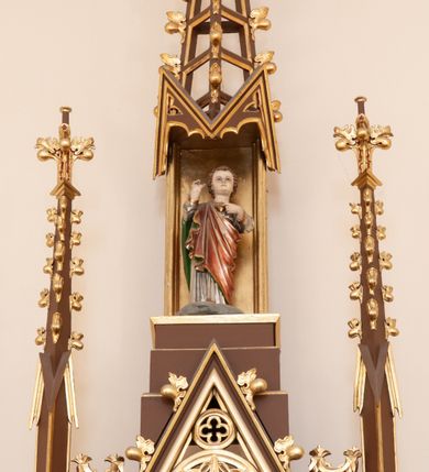 Zdjęcie nr 7: Ołtarz przyścienny, architektoniczny, pięcioosiowy, jednokondygnacyjny ze zwieńczeniem. Antepedium dekorowane płyciną w profilowanej ramie z umieszczonym centralnie czteroliściem oraz krzyżem. Tabernakulum metalowe, jednodrzwiczkowe z umieszczonym centralnie kielichem z hostią na tle kłosów oraz winorośli. Po bokach umieszczone litery „A” i „Ω” na tle krzyża każda. Nastawa posadowiona na wysokim cokole z płycinami wypełnionymi maswerkiem. Osie w układzie schodkowym z osią środkową wyższą i szerszą od bocznych, wydzielone wiązkami służek na postumentach w formie skręconych trzonów, z kapitelami dekorowanymi motywem ostroliścia. Służki zwieńczone pinaklami zakończonymi kwiatonami, w osi środkowej ażurowa wieżyczka w formie zbliżonej do pinakla zakończonego kwiatonem i krzyżem, tworząca baldachim nad figurą Dzieciątka Jezus. Osie środkowe zwieńczone trójkątnymi wimpergami wypełnionymi dekoracją maswerkową. W polu ołtarza obraz Trójcy Świętej w złoconej, profilowanej ramie w kształcie stojącego prostokąta zamkniętego łukiem ostrym z maswerkiem. Poniżej płycina z płaskorzeźbionym krzyżem na tle kłosów. W osiach drugiej i czwartej złocone nisze mieszczące figury św. Stanisława i Wojciecha. Nisze zamknięte od góry muszlami palmetowymi. Pod niszami płyciny wypełnione maswerkiem z fantazyjnym motywem geometrycznym. Osie skrajne wypełnione zdwojonymi blendami z rozetami pośrodku. Struktura ołtarza malowana na brązowo z elementami czerwieni, detal architektoniczny złocony. Figury w pełni polichromowane.