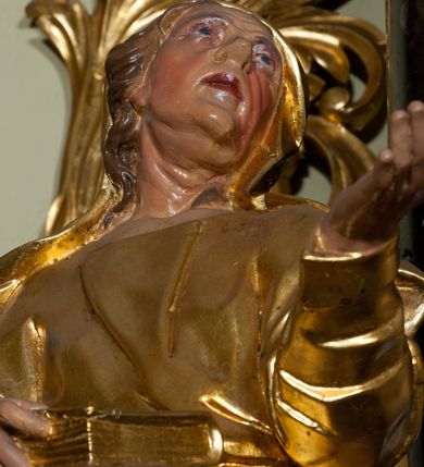 Zdjęcie nr 2: Rzeźba ścięta z tyłu, drążona, ustawiona na niskiej podstawie. Figura stojąca, w kontrapoście, zwrócona frontalnie z głową przechyloną na lewe ramię, skierowaną ku górze. Jej lewa ręka jest wyciągnięta przed siebie, a w prawej dłoni trzyma księgę opartą na biodrze. Twarz ascetyczna, o starczych rysach, głęboko osadzone oczy z silnie uwypuklonymi brwiami, nos niewielki, usta lekko rozchylone, podbródek wydatny. Włosy długie, delikatnie kręcone, opadają na ramiona. Święta jest ubrana w suknię o wąskich, drapowanych rękawach, przepasaną w talii oraz płaszcz, przewiązany w partii bioder, opadający na prawy bok, długi, odsłaniający fragmenty stóp. Na głowie ściśle do niej przylegający welon. Polichromia naturalistyczna w odsłoniętych partiach ciała. Szaty i atrybut złocony.