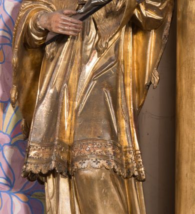 Zdjęcie nr 2: Rzeźba ścięta z tyłu, drążona, ustawiona na prostopadłościennym, niskim cokole. Święty Jan Nepomucen ukazany w lekkim kontrapoście, zwrócony frontalnie, z głową delikatnie pochyloną. Postać obiema rękami podtrzymuje krucyfiks. Jego twarz jest ascetyczna, o uwypuklonej linii łuków brwiowych, kości policzkowych i szpiczastej brodzie. Oczy ma głęboko osadzone, lekko przymknięte, spoglądające w dół, nos szeroki, wydatny, usta pełne, delikatnie rozchylone. Twarz okolona krótką, kręconą, szpiczastą brodą, rozdwojoną na końcu. Włosy krótkie, układające się w odstające pukle, lekko kręcone, czarne. Święty jest ubrany w albę, drapowaną w równoległe fałdy, komżę z rytą koronką u brzegu, almucję z chwostami, zawiązaną u szyi. Polichromia naturalistyczna w odsłoniętych partiach ciała; szaty złocone.