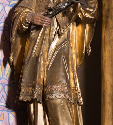 Zdjęcie nr 3: Rzeźba ścięta z tyłu, drążona, ustawiona na prostopadłościennym, niskim cokole. Święty Jan Nepomucen ukazany w lekkim kontrapoście, zwrócony frontalnie, z głową delikatnie pochyloną. Postać obiema rękami podtrzymuje krucyfiks. Jego twarz jest ascetyczna, o uwypuklonej linii łuków brwiowych, kości policzkowych i szpiczastej brodzie. Oczy ma głęboko osadzone, lekko przymknięte, spoglądające w dół, nos szeroki, wydatny, usta pełne, delikatnie rozchylone. Twarz okolona krótką, kręconą, szpiczastą brodą, rozdwojoną na końcu. Włosy krótkie, układające się w odstające pukle, lekko kręcone, czarne. Święty jest ubrany w albę, drapowaną w równoległe fałdy, komżę z rytą koronką u brzegu, almucję z chwostami, zawiązaną u szyi. Polichromia naturalistyczna w odsłoniętych partiach ciała; szaty złocone.