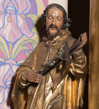 Zdjęcie nr 1: Rzeźba ścięta z tyłu, drążona, ustawiona na prostopadłościennym, niskim cokole. Święty Jan Nepomucen ukazany w lekkim kontrapoście, zwrócony frontalnie, z głową delikatnie pochyloną. Postać obiema rękami podtrzymuje krucyfiks. Jego twarz jest ascetyczna, o uwypuklonej linii łuków brwiowych, kości policzkowych i szpiczastej brodzie. Oczy ma głęboko osadzone, lekko przymknięte, spoglądające w dół, nos szeroki, wydatny, usta pełne, delikatnie rozchylone. Twarz okolona krótką, kręconą, szpiczastą brodą, rozdwojoną na końcu. Włosy krótkie, układające się w odstające pukle, lekko kręcone, czarne. Święty jest ubrany w albę, drapowaną w równoległe fałdy, komżę z rytą koronką u brzegu, almucję z chwostami, zawiązaną u szyi. Polichromia naturalistyczna w odsłoniętych partiach ciała; szaty złocone.