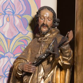 Zdjęcie nr 1: Rzeźba ścięta z tyłu, drążona, ustawiona na prostopadłościennym, niskim cokole. Święty Jan Nepomucen ukazany w lekkim kontrapoście, zwrócony frontalnie, z głową delikatnie pochyloną. Postać obiema rękami podtrzymuje krucyfiks. Jego twarz jest ascetyczna, o uwypuklonej linii łuków brwiowych, kości policzkowych i szpiczastej brodzie. Oczy ma głęboko osadzone, lekko przymknięte, spoglądające w dół, nos szeroki, wydatny, usta pełne, delikatnie rozchylone. Twarz okolona krótką, kręconą, szpiczastą brodą, rozdwojoną na końcu. Włosy krótkie, układające się w odstające pukle, lekko kręcone, czarne. Święty jest ubrany w albę, drapowaną w równoległe fałdy, komżę z rytą koronką u brzegu, almucję z chwostami, zawiązaną u szyi. Polichromia naturalistyczna w odsłoniętych partiach ciała; szaty złocone.