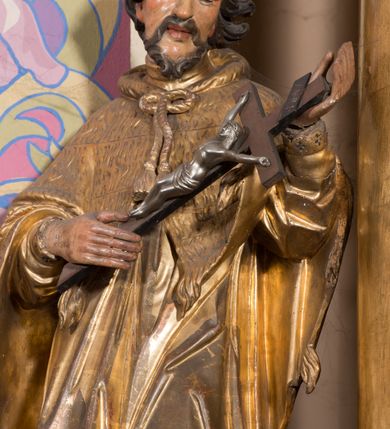 Zdjęcie nr 4: Rzeźba ścięta z tyłu, drążona, ustawiona na prostopadłościennym, niskim cokole. Święty Jan Nepomucen ukazany w lekkim kontrapoście, zwrócony frontalnie, z głową delikatnie pochyloną. Postać obiema rękami podtrzymuje krucyfiks. Jego twarz jest ascetyczna, o uwypuklonej linii łuków brwiowych, kości policzkowych i szpiczastej brodzie. Oczy ma głęboko osadzone, lekko przymknięte, spoglądające w dół, nos szeroki, wydatny, usta pełne, delikatnie rozchylone. Twarz okolona krótką, kręconą, szpiczastą brodą, rozdwojoną na końcu. Włosy krótkie, układające się w odstające pukle, lekko kręcone, czarne. Święty jest ubrany w albę, drapowaną w równoległe fałdy, komżę z rytą koronką u brzegu, almucję z chwostami, zawiązaną u szyi. Polichromia naturalistyczna w odsłoniętych partiach ciała; szaty złocone.