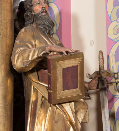 Zdjęcie nr 6: Rzeźba ścięta z tyłu, drążona, ustawiona na cokole w formie kostki. Figura ustawiona w kontrapoście, zwrócona frontalnie, o esowato wygiętej sylwetce. W lewej, odsuniętej od boku ręce trzyma miecz, zwrócony ostrzem w dół, prawą podtrzymuje na udzie zamkniętą księgę. Twarz ma pociągłą, szczupłą, oczy w głębokich oczodołach z silnie zaznaczonymi i podniesionymi łukami brwiowymi, nos mały, usta pełne. Jego twarz jest okolona długą brodą opadającą lokami na pierś z rozdwojonymi końcówkami. Włosy kręcone, głęboko rzeźbione, sięgające szyi. Święty jest ubrany w tunikę o wąskich, drapowanych rękawach, ściśle przylegającą do ciała, przepasaną w talii szarfą oraz obficie drapowany i łamany płaszcz przewiązany przez lewe ramię i opadający na prawą nogę. Polichromia naturalistyczna w odsłoniętych partiach ciała, szaty złocone, atrybuty złocone i srebrzone.