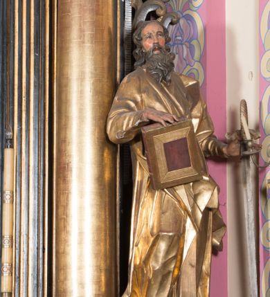 Zdjęcie nr 1: Rzeźba ścięta z tyłu, drążona, ustawiona na cokole w formie kostki. Figura ustawiona w kontrapoście, zwrócona frontalnie, o esowato wygiętej sylwetce. W lewej, odsuniętej od boku ręce trzyma miecz, zwrócony ostrzem w dół, prawą podtrzymuje na udzie zamkniętą księgę. Twarz ma pociągłą, szczupłą, oczy w głębokich oczodołach z silnie zaznaczonymi i podniesionymi łukami brwiowymi, nos mały, usta pełne. Jego twarz jest okolona długą brodą opadającą lokami na pierś z rozdwojonymi końcówkami. Włosy kręcone, głęboko rzeźbione, sięgające szyi. Święty jest ubrany w tunikę o wąskich, drapowanych rękawach, ściśle przylegającą do ciała, przepasaną w talii szarfą oraz obficie drapowany i łamany płaszcz przewiązany przez lewe ramię i opadający na prawą nogę. Polichromia naturalistyczna w odsłoniętych partiach ciała, szaty złocone, atrybuty złocone i srebrzone.