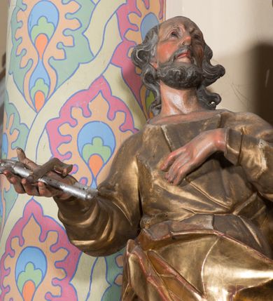 Zdjęcie nr 4: Rzeźba ścięta z tyłu, drążona, ustawiona na prostokątnej podstawie. Figura zwrócona frontalnie, z głową skierowaną w lewo, uniesiona do góry. Ciało ukazane w kontrapoście, przechylone w tył. Święty lewą rękę kładzie na piersi, w prawej wysuniętej do przodu trzyma skrzyżowane klucze. Jego twarz jest ascetyczna o ostrych rysach, głęboko osadzonych oczach o trójkątnych, wysoko uniesionych powiekach, nos ma niewielki, okrągły, a usta delikatnie rozchylone z widocznym podniebieniem. Twarz okolona krótką brodą, układającą się w dwa pukle. Włosy kręcone, głęboko rzeźbione, opadające na tył szyi. Święty jest ubrany w tunikę o wąskich drapowanych rękawach, ściśle przylegającą do ciała w partii torsu oraz obficie drapowaną i delikatnie łamaną w dolnej części. Płaszcz przewiązany przez biodra z rozwianym zwisem na prawym boku. Polichromia naturalistyczna w odsłoniętych partiach ciała, szaty złocone, klucze srebrzone.