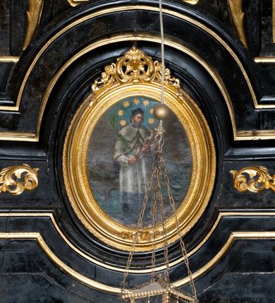 Zdjęcie nr 2: Obraz w formie stojącego owalu, w drewnianej, złoconej, profilowanej ramie, u szczytu dekorowanej ornamentem roślinnym. W centrum przedstawienie stojącego św. Jana Nepomucena, zwróconego w trzech czwartych w lewo, z krucyfiksem i palmą męczeństwa w dłoniach. Jego głowa jest delikatnie pochylona, twarz okolona krótką brodą, włosy krótkie. Jest ubrany w sutannę i komżę, almucję zapinaną na piersi, opadającą na ramiona oraz biret na głowie. Wokół głowy świetlisty nimb z siedmioma gwiazdami. Święty jest przedstawiony na tle mostu Karola w Pradze i architektonicznego pejzażu miejskiego, nad którym szaro-niebieskie niebo. 