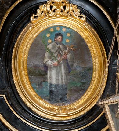 Zdjęcie nr 3: Obraz w formie stojącego owalu, w drewnianej, złoconej, profilowanej ramie, u szczytu dekorowanej ornamentem roślinnym. W centrum przedstawienie stojącego św. Jana Nepomucena, zwróconego w trzech czwartych w lewo, z krucyfiksem i palmą męczeństwa w dłoniach. Jego głowa jest delikatnie pochylona, twarz okolona krótką brodą, włosy krótkie. Jest ubrany w sutannę i komżę, almucję zapinaną na piersi, opadającą na ramiona oraz biret na głowie. Wokół głowy świetlisty nimb z siedmioma gwiazdami. Święty jest przedstawiony na tle mostu Karola w Pradze i architektonicznego pejzażu miejskiego, nad którym szaro-niebieskie niebo. 