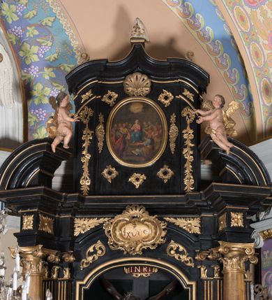 Zdjęcie nr 2: Ołtarz architektoniczny, jednoosiowy, jednokondygnacyjny ze zwieńczeniem. Ołtarz drewniany prostopadłościenny, mensa drewniana z portatylem z marmuru dębnickiego. W ołtarzu nisza z Grobem Pańskim, zakryta antepedium dekorowanym ramą w formie leżącego prostokąta o wklęsłych narożach z monogramem IHS w polu. Na ściankach bocznych ołtarza płyciny w formie kwadratu o wklęsłych narożach. Retabulum jest ujęte parą kompozytowych kolumn ustawionych na ukośnych cokołach stojących, na tle zwielokrotnionych pilastrów o kanelowanych trzonach, dźwigających pełne belkowanie z wydatnym gzymsem wyłamane na osi podpór oraz przerwany półkolisty przyczółek z figurami siedzących puttów; architraw nad polem środkowym przybiera kształt łuku odcinkowego. Po zewnętrznej stronie kolumn figury św. Piotra i św. Pawła stojące na wydatnych konsolach. W polu środkowym nisza ujęta profilowaną ramą w formie stojącego prostokąta zamkniętego łukiem odcinkowym nadwieszonym z figurą Chrystusa Ukrzyżowanego. Zwieńczenie w formie stojącego prostokąta ujęte zwielokrotnionymi pilastrami podtrzymującymi profilowany gzyms przyjmujący kształt łuku oślego. W polu owalne pole z obrazem „Zesłanie Ducha Świętego”. Cokoły dekorowane płycinami utworzonymi z ornamentu roślinnego, zaś konsole zdobione muszlą o kształcie falbany. Nad polem środkowym rocaille'owy kartusz z napisem: „IXEUS”, flankowany akantem. U spodu obrazu prostokątna płycina. Fryz zdobiony rozetami. Kondygnacja ujęta ażurowymi uszami utworzonymi z suchego akantu, wstęgi, rocaille'u i rozet. Trzony pilastrów zwieńczenia dekorowane rocaille'em i roślinnymi festonami. Pole obrazowe otoczone fragmentami rocaille'u, zwieńczone muszlą. Ponad zwieńczeniem krzyż lub wazon nakryty sukienką na puszkę. Struktura polichromowana w kolorze czarnym; kolumny, ornamentyka i profile złocone.