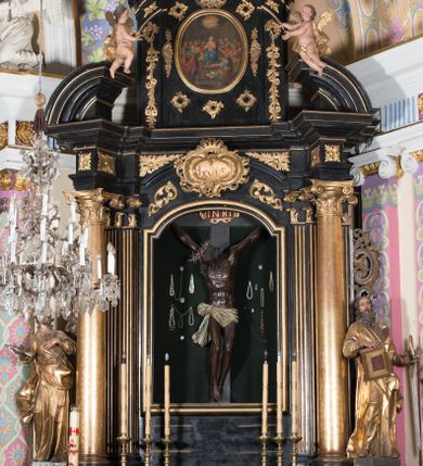 Zdjęcie nr 1: Ołtarz architektoniczny, jednoosiowy, jednokondygnacyjny ze zwieńczeniem. Ołtarz drewniany prostopadłościenny, mensa drewniana z portatylem z marmuru dębnickiego. W ołtarzu nisza z Grobem Pańskim, zakryta antepedium dekorowanym ramą w formie leżącego prostokąta o wklęsłych narożach z monogramem IHS w polu. Na ściankach bocznych ołtarza płyciny w formie kwadratu o wklęsłych narożach. Retabulum jest ujęte parą kompozytowych kolumn ustawionych na ukośnych cokołach stojących, na tle zwielokrotnionych pilastrów o kanelowanych trzonach, dźwigających pełne belkowanie z wydatnym gzymsem wyłamane na osi podpór oraz przerwany półkolisty przyczółek z figurami siedzących puttów; architraw nad polem środkowym przybiera kształt łuku odcinkowego. Po zewnętrznej stronie kolumn figury św. Piotra i św. Pawła stojące na wydatnych konsolach. W polu środkowym nisza ujęta profilowaną ramą w formie stojącego prostokąta zamkniętego łukiem odcinkowym nadwieszonym z figurą Chrystusa Ukrzyżowanego. Zwieńczenie w formie stojącego prostokąta ujęte zwielokrotnionymi pilastrami podtrzymującymi profilowany gzyms przyjmujący kształt łuku oślego. W polu owalne pole z obrazem „Zesłanie Ducha Świętego”. Cokoły dekorowane płycinami utworzonymi z ornamentu roślinnego, zaś konsole zdobione muszlą o kształcie falbany. Nad polem środkowym rocaille'owy kartusz z napisem: „IXEUS”, flankowany akantem. U spodu obrazu prostokątna płycina. Fryz zdobiony rozetami. Kondygnacja ujęta ażurowymi uszami utworzonymi z suchego akantu, wstęgi, rocaille'u i rozet. Trzony pilastrów zwieńczenia dekorowane rocaille'em i roślinnymi festonami. Pole obrazowe otoczone fragmentami rocaille'u, zwieńczone muszlą. Ponad zwieńczeniem krzyż lub wazon nakryty sukienką na puszkę. Struktura polichromowana w kolorze czarnym; kolumny, ornamentyka i profile złocone.