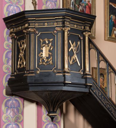 Zdjęcie nr 2: Ambona dostępna schodami od wschodniej strony w korpusie nawowym. Kosz na rzucie sześcioboku, wydzielony profilowanym belkowaniem. Podziały pionowe pól kosza wyznaczone naprzemiennie parami pilastrów o kanelowanych trzonach i pólkolumienek z jońskimi kapitelami. Pola kosza zdobione płycinami w kształcie stojącego prostokąta o wklęsło ściętych  narożach. Płyciny zdobione ornamentyką roślinną, dodatkowo w pierwszej od wschodu potłuczone tablice Dekalogu, na drugiej motyw krzyża, kotwicy i serca Jezusa, na trzeciej dwie tablice Mojżeszowe. Fryz zdobi motyw tryglifu na przemian z rozetkami. Schodki z drewnianą balustradą z motywem arkadki i ornamentyki roślinnej, w dolnej części niewielka bramka zdobiona prostą ornamentyka roślinną. Struktura polichromowana na czarno, partie detalu architektonicznego i ornamentyka złocone.