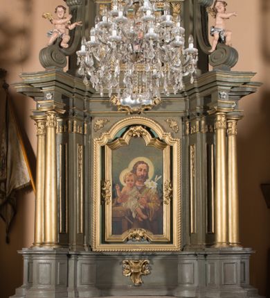 Zdjęcie nr 1: Nastawa architektoniczna, jednokondygnacyjna, jednoosiowa ze zwieńczeniem. Ołtarz sarkofagowy, wybrzuszony, z drewnianą mensą i drewnianym portatylem integralnie zespolony z cokołem retabulum. Nastawa wsparta na ukośnie ustawionych postumentach z filarami i stojącymi przed nimi oraz z boku wolnostojącymi kolumnami z głowicami akantowymi, podtrzymującymi wspólnie, wznoszące się nad nimi belkowanie. Zwieńczenie ujęte ukośnie posadowionymi spływami wolutowymi podtrzymującymi gzyms wyłamujący się pośrodku łukowato. W środku nastawy głęboka wnęka w profilowanym obramieniu z motywem skręconego sznura, prostokątna o ściętych górą narożach, zamknięta łukiem półkolistym nadwieszonym. W niej obraz przedstawiający św. Józefa z Dzieciątkiem, w ramie powielającej wykrój wnęki, z drobnymi rocaille'ami na bokach. W zwieńczeniu wnęka prostokątna w profilowanym obramieniu, z motywem skręconego sznura, ujęta po bokach kłosami zbóż, a zwieńczona motywem akantu. W niej obraz prostokątny obraz z wyobrażeniem św. Stanisława Kostki. Na spływach wolutowych uskrzydlone nagie aniołki z przepaską na biodrach. Nad wnęką w środkowej części ołtarza wypukły kartusz ujęty rocaile'm, drobne rocaille nad wnęką i na filarach, w partii cokołu utworzona z motywu rocaille'owego konsola.  Kolorystyka  struktury w odcieniu  zieleni wpadającej w szarość, partie detalu architektonicznego i ornamentyka  złocone.