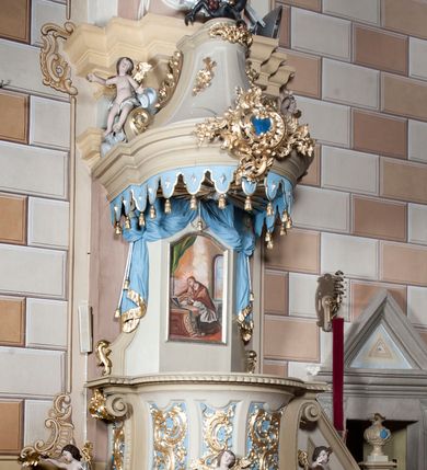 Zdjęcie nr 2: Ambona nadwieszona z dostępem w postaci jednobiegowych schodów usytuowanych z prawej strony kosza. Kosz na rzucie półkolistym, z podwieszoną szyszką u spodu. Ścianki kosza flankowane dwoma spływami wolutowymi. Cokół kosza zaakcentowany szeregiem form przypominających obłoki. Ścianki kosza podzielone na sześć  pionowych, prostokątnych, o wciętych półkoliście krótszych bokach, wypełnionych dekoracją z motywów rocaille. Gzyms profilowany, przełamany na osi spływów. Na wolutach spływów oraz pośrodku kosza osadzone trzy figury aniołków. Zaplecek rozszerzający się ku dołowi, flankowany parą spływów; w jego polu obraz w kształcie pionowego prostokąta zamkniętego łukiem wklęsło-wypukłym z przedstawieniem św. Jana Nepomucena. Zaplecek ujęty z boków i zwieńczony spiętą kotarą z frędzlami. Baldachim na planie analogicznym jak kosz, o wysokim, profilowanym dolnym brzegu, obwiedziony od dołu lambrekinem z frędzlami. W polu podniebienia baldachimu gołębica Ducha Świętego na tle promienistej glorii, z przodu na brzegu baldachimu kartusz z motywu rocaille. Podniebienie baldachimu w kolorze niebieskim, na polu gołębica Ducha Świętego na tle promienistej glorii, z przodu rocaille'owy kartusz. Szczyt baldachimu w formie wysokiego postumentu, na którym ustawiona rzeźba Michała Archanioła zwyciężającego szatana. Po bokach postumentu dwa rzeźbione aniołki. Święty Michał Archanioł ukazany w pozycji stojącej o esowato wygiętej sylwetce, z prawą ręką uniesioną wysoko ku górze, w której trzyma płomienisty miecz, w lewej owalna tarcza z monogramem Chrystusa i trzema gwoździami, lewą stopą depcze leżącego szatana. Ubrany w strój legionisty rzymskiego, skrzydła złocone, szeroko rozłożone. Szatan ukazany w monstrualnej postaci, w pozycji leżącej na brzuchu, z lewą nogą uniesioną wysoko ku górze. Postać polichromowana na czarno, detale (oczy, usta, rogi, paznokcie) na czerwono. Aniołki ukazane w poruszonych pozach, przepasane srebrną chustką, z szeroko rozłożonymi złoconymi skrzydłami. Balustrada schodów zamknięta od dołu cokołem i od góry profilowanym gzymsem, zdobiona w polu romboidalną płyciną, zakończona słupkiem z wazonem płomienistym na szczycie. Struktura ambony polichromowana na kolor jasno i ciemnobeżowy, detale i płyciny na kolor niebieski, ornamentyka złocona.
