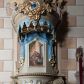 Zdjęcie nr 1: Ambona nadwieszona z dostępem w postaci jednobiegowych schodów usytuowanych z prawej strony kosza. Kosz na rzucie półkolistym, z podwieszoną szyszką u spodu. Ścianki kosza flankowane dwoma spływami wolutowymi. Cokół kosza zaakcentowany szeregiem form przypominających obłoki. Ścianki kosza podzielone na sześć  pionowych, prostokątnych, o wciętych półkoliście krótszych bokach, wypełnionych dekoracją z motywów rocaille. Gzyms profilowany, przełamany na osi spływów. Na wolutach spływów oraz pośrodku kosza osadzone trzy figury aniołków. Zaplecek rozszerzający się ku dołowi, flankowany parą spływów; w jego polu obraz w kształcie pionowego prostokąta zamkniętego łukiem wklęsło-wypukłym z przedstawieniem św. Jana Nepomucena. Zaplecek ujęty z boków i zwieńczony spiętą kotarą z frędzlami. Baldachim na planie analogicznym jak kosz, o wysokim, profilowanym dolnym brzegu, obwiedziony od dołu lambrekinem z frędzlami. W polu podniebienia baldachimu gołębica Ducha Świętego na tle promienistej glorii, z przodu na brzegu baldachimu kartusz z motywu rocaille. Podniebienie baldachimu w kolorze niebieskim, na polu gołębica Ducha Świętego na tle promienistej glorii, z przodu rocaille'owy kartusz. Szczyt baldachimu w formie wysokiego postumentu, na którym ustawiona rzeźba Michała Archanioła zwyciężającego szatana. Po bokach postumentu dwa rzeźbione aniołki. Święty Michał Archanioł ukazany w pozycji stojącej o esowato wygiętej sylwetce, z prawą ręką uniesioną wysoko ku górze, w której trzyma płomienisty miecz, w lewej owalna tarcza z monogramem Chrystusa i trzema gwoździami, lewą stopą depcze leżącego szatana. Ubrany w strój legionisty rzymskiego, skrzydła złocone, szeroko rozłożone. Szatan ukazany w monstrualnej postaci, w pozycji leżącej na brzuchu, z lewą nogą uniesioną wysoko ku górze. Postać polichromowana na czarno, detale (oczy, usta, rogi, paznokcie) na czerwono. Aniołki ukazane w poruszonych pozach, przepasane srebrną chustką, z szeroko rozłożonymi złoconymi skrzydłami. Balustrada schodów zamknięta od dołu cokołem i od góry profilowanym gzymsem, zdobiona w polu romboidalną płyciną, zakończona słupkiem z wazonem płomienistym na szczycie. Struktura ambony polichromowana na kolor jasno i ciemnobeżowy, detale i płyciny na kolor niebieski, ornamentyka złocona.
