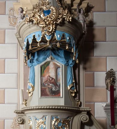 Zdjęcie nr 1: Ambona nadwieszona z dostępem w postaci jednobiegowych schodów usytuowanych z prawej strony kosza. Kosz na rzucie półkolistym, z podwieszoną szyszką u spodu. Ścianki kosza flankowane dwoma spływami wolutowymi. Cokół kosza zaakcentowany szeregiem form przypominających obłoki. Ścianki kosza podzielone na sześć  pionowych, prostokątnych, o wciętych półkoliście krótszych bokach, wypełnionych dekoracją z motywów rocaille. Gzyms profilowany, przełamany na osi spływów. Na wolutach spływów oraz pośrodku kosza osadzone trzy figury aniołków. Zaplecek rozszerzający się ku dołowi, flankowany parą spływów; w jego polu obraz w kształcie pionowego prostokąta zamkniętego łukiem wklęsło-wypukłym z przedstawieniem św. Jana Nepomucena. Zaplecek ujęty z boków i zwieńczony spiętą kotarą z frędzlami. Baldachim na planie analogicznym jak kosz, o wysokim, profilowanym dolnym brzegu, obwiedziony od dołu lambrekinem z frędzlami. W polu podniebienia baldachimu gołębica Ducha Świętego na tle promienistej glorii, z przodu na brzegu baldachimu kartusz z motywu rocaille. Podniebienie baldachimu w kolorze niebieskim, na polu gołębica Ducha Świętego na tle promienistej glorii, z przodu rocaille'owy kartusz. Szczyt baldachimu w formie wysokiego postumentu, na którym ustawiona rzeźba Michała Archanioła zwyciężającego szatana. Po bokach postumentu dwa rzeźbione aniołki. Święty Michał Archanioł ukazany w pozycji stojącej o esowato wygiętej sylwetce, z prawą ręką uniesioną wysoko ku górze, w której trzyma płomienisty miecz, w lewej owalna tarcza z monogramem Chrystusa i trzema gwoździami, lewą stopą depcze leżącego szatana. Ubrany w strój legionisty rzymskiego, skrzydła złocone, szeroko rozłożone. Szatan ukazany w monstrualnej postaci, w pozycji leżącej na brzuchu, z lewą nogą uniesioną wysoko ku górze. Postać polichromowana na czarno, detale (oczy, usta, rogi, paznokcie) na czerwono. Aniołki ukazane w poruszonych pozach, przepasane srebrną chustką, z szeroko rozłożonymi złoconymi skrzydłami. Balustrada schodów zamknięta od dołu cokołem i od góry profilowanym gzymsem, zdobiona w polu romboidalną płyciną, zakończona słupkiem z wazonem płomienistym na szczycie. Struktura ambony polichromowana na kolor jasno i ciemnobeżowy, detale i płyciny na kolor niebieski, ornamentyka złocona.
