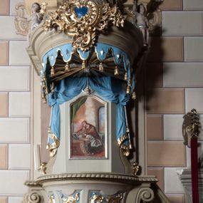 Zdjęcie nr 1: Ambona nadwieszona z dostępem w postaci jednobiegowych schodów usytuowanych z prawej strony kosza. Kosz na rzucie półkolistym, z podwieszoną szyszką u spodu. Ścianki kosza flankowane dwoma spływami wolutowymi. Cokół kosza zaakcentowany szeregiem form przypominających obłoki. Ścianki kosza podzielone na sześć  pionowych, prostokątnych, o wciętych półkoliście krótszych bokach, wypełnionych dekoracją z motywów rocaille. Gzyms profilowany, przełamany na osi spływów. Na wolutach spływów oraz pośrodku kosza osadzone trzy figury aniołków. Zaplecek rozszerzający się ku dołowi, flankowany parą spływów; w jego polu obraz w kształcie pionowego prostokąta zamkniętego łukiem wklęsło-wypukłym z przedstawieniem św. Jana Nepomucena. Zaplecek ujęty z boków i zwieńczony spiętą kotarą z frędzlami. Baldachim na planie analogicznym jak kosz, o wysokim, profilowanym dolnym brzegu, obwiedziony od dołu lambrekinem z frędzlami. W polu podniebienia baldachimu gołębica Ducha Świętego na tle promienistej glorii, z przodu na brzegu baldachimu kartusz z motywu rocaille. Podniebienie baldachimu w kolorze niebieskim, na polu gołębica Ducha Świętego na tle promienistej glorii, z przodu rocaille'owy kartusz. Szczyt baldachimu w formie wysokiego postumentu, na którym ustawiona rzeźba Michała Archanioła zwyciężającego szatana. Po bokach postumentu dwa rzeźbione aniołki. Święty Michał Archanioł ukazany w pozycji stojącej o esowato wygiętej sylwetce, z prawą ręką uniesioną wysoko ku górze, w której trzyma płomienisty miecz, w lewej owalna tarcza z monogramem Chrystusa i trzema gwoździami, lewą stopą depcze leżącego szatana. Ubrany w strój legionisty rzymskiego, skrzydła złocone, szeroko rozłożone. Szatan ukazany w monstrualnej postaci, w pozycji leżącej na brzuchu, z lewą nogą uniesioną wysoko ku górze. Postać polichromowana na czarno, detale (oczy, usta, rogi, paznokcie) na czerwono. Aniołki ukazane w poruszonych pozach, przepasane srebrną chustką, z szeroko rozłożonymi złoconymi skrzydłami. Balustrada schodów zamknięta od dołu cokołem i od góry profilowanym gzymsem, zdobiona w polu romboidalną płyciną, zakończona słupkiem z wazonem płomienistym na szczycie. Struktura ambony polichromowana na kolor jasno i ciemnobeżowy, detale i płyciny na kolor niebieski, ornamentyka złocona.
