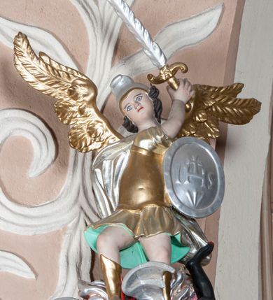 Zdjęcie nr 9: Ambona nadwieszona z dostępem w postaci jednobiegowych schodów usytuowanych z prawej strony kosza. Kosz na rzucie półkolistym, z podwieszoną szyszką u spodu. Ścianki kosza flankowane dwoma spływami wolutowymi. Cokół kosza zaakcentowany szeregiem form przypominających obłoki. Ścianki kosza podzielone na sześć  pionowych, prostokątnych, o wciętych półkoliście krótszych bokach, wypełnionych dekoracją z motywów rocaille. Gzyms profilowany, przełamany na osi spływów. Na wolutach spływów oraz pośrodku kosza osadzone trzy figury aniołków. Zaplecek rozszerzający się ku dołowi, flankowany parą spływów; w jego polu obraz w kształcie pionowego prostokąta zamkniętego łukiem wklęsło-wypukłym z przedstawieniem św. Jana Nepomucena. Zaplecek ujęty z boków i zwieńczony spiętą kotarą z frędzlami. Baldachim na planie analogicznym jak kosz, o wysokim, profilowanym dolnym brzegu, obwiedziony od dołu lambrekinem z frędzlami. W polu podniebienia baldachimu gołębica Ducha Świętego na tle promienistej glorii, z przodu na brzegu baldachimu kartusz z motywu rocaille. Podniebienie baldachimu w kolorze niebieskim, na polu gołębica Ducha Świętego na tle promienistej glorii, z przodu rocaille'owy kartusz. Szczyt baldachimu w formie wysokiego postumentu, na którym ustawiona rzeźba Michała Archanioła zwyciężającego szatana. Po bokach postumentu dwa rzeźbione aniołki. Święty Michał Archanioł ukazany w pozycji stojącej o esowato wygiętej sylwetce, z prawą ręką uniesioną wysoko ku górze, w której trzyma płomienisty miecz, w lewej owalna tarcza z monogramem Chrystusa i trzema gwoździami, lewą stopą depcze leżącego szatana. Ubrany w strój legionisty rzymskiego, skrzydła złocone, szeroko rozłożone. Szatan ukazany w monstrualnej postaci, w pozycji leżącej na brzuchu, z lewą nogą uniesioną wysoko ku górze. Postać polichromowana na czarno, detale (oczy, usta, rogi, paznokcie) na czerwono. Aniołki ukazane w poruszonych pozach, przepasane srebrną chustką, z szeroko rozłożonymi złoconymi skrzydłami. Balustrada schodów zamknięta od dołu cokołem i od góry profilowanym gzymsem, zdobiona w polu romboidalną płyciną, zakończona słupkiem z wazonem płomienistym na szczycie. Struktura ambony polichromowana na kolor jasno i ciemnobeżowy, detale i płyciny na kolor niebieski, ornamentyka złocona.
