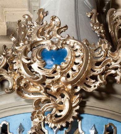 Zdjęcie nr 3: Ambona nadwieszona z dostępem w postaci jednobiegowych schodów usytuowanych z prawej strony kosza. Kosz na rzucie półkolistym, z podwieszoną szyszką u spodu. Ścianki kosza flankowane dwoma spływami wolutowymi. Cokół kosza zaakcentowany szeregiem form przypominających obłoki. Ścianki kosza podzielone na sześć  pionowych, prostokątnych, o wciętych półkoliście krótszych bokach, wypełnionych dekoracją z motywów rocaille. Gzyms profilowany, przełamany na osi spływów. Na wolutach spływów oraz pośrodku kosza osadzone trzy figury aniołków. Zaplecek rozszerzający się ku dołowi, flankowany parą spływów; w jego polu obraz w kształcie pionowego prostokąta zamkniętego łukiem wklęsło-wypukłym z przedstawieniem św. Jana Nepomucena. Zaplecek ujęty z boków i zwieńczony spiętą kotarą z frędzlami. Baldachim na planie analogicznym jak kosz, o wysokim, profilowanym dolnym brzegu, obwiedziony od dołu lambrekinem z frędzlami. W polu podniebienia baldachimu gołębica Ducha Świętego na tle promienistej glorii, z przodu na brzegu baldachimu kartusz z motywu rocaille. Podniebienie baldachimu w kolorze niebieskim, na polu gołębica Ducha Świętego na tle promienistej glorii, z przodu rocaille'owy kartusz. Szczyt baldachimu w formie wysokiego postumentu, na którym ustawiona rzeźba Michała Archanioła zwyciężającego szatana. Po bokach postumentu dwa rzeźbione aniołki. Święty Michał Archanioł ukazany w pozycji stojącej o esowato wygiętej sylwetce, z prawą ręką uniesioną wysoko ku górze, w której trzyma płomienisty miecz, w lewej owalna tarcza z monogramem Chrystusa i trzema gwoździami, lewą stopą depcze leżącego szatana. Ubrany w strój legionisty rzymskiego, skrzydła złocone, szeroko rozłożone. Szatan ukazany w monstrualnej postaci, w pozycji leżącej na brzuchu, z lewą nogą uniesioną wysoko ku górze. Postać polichromowana na czarno, detale (oczy, usta, rogi, paznokcie) na czerwono. Aniołki ukazane w poruszonych pozach, przepasane srebrną chustką, z szeroko rozłożonymi złoconymi skrzydłami. Balustrada schodów zamknięta od dołu cokołem i od góry profilowanym gzymsem, zdobiona w polu romboidalną płyciną, zakończona słupkiem z wazonem płomienistym na szczycie. Struktura ambony polichromowana na kolor jasno i ciemnobeżowy, detale i płyciny na kolor niebieski, ornamentyka złocona.
