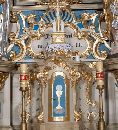 Zdjęcie nr 25: Nastawa ołtarzowa architektoniczna, jednoosiowa, jednokondygnacyjna ze zwieńczeniem, z bramkami i z rocaille'owymi uszami. Tabernakulum w formie zbliżonej do prostopadłościennej skrzynki, na wklęsło-wypukłym rzucie, ustawione na niskim cokole, o wypukłej ściance frontowej. Ujęte jest drewnianą obudową o wklęsłych bokach, flankowane parą ustawionych pod kątem pilastrów, których trzony przyjmują formę wolut. Zwieńczenie tabernakulum w formie niewielkiego szczytu, na którym ustawiona jest pasyjka. Całość ujęta po bokach rocaille'owymi wolutami. Drzwiczki w kształcie stojącego prostokąta, zamknięte łukiem nadwieszonym. W polu, na niebieskim tle malowane przedstawienie kielicha z hostią. Nastawa ustawiona na zdwojonym cokole, o  wklęsłym przebiegu, ujęta dwiema parami kolumn na tle zwielokrotnionych pilastrów. Podpory dźwigają pełne belkowanie, wyłamane pośrodku. Pomiędzy podporami figury: po lewej św. Józefa, a po prawej św. Jana Chrzciciela, nad bramkami: po lewej św. Wojciecha, a po prawej św. Stanisława. Nad tabernakulum rocaille'owy kartusz z łacińską inskrypcją w polu. W polu środkowym nisza w formie stojącego prostokąta, zamkniętego łukiem wklęsło-wypukłym z uskokami z obrazem „Święta Rozmowa”. Zwieńczenie w formie zbliżonej do stojącego prostokąta ujętego po bokach dwoma pilastrami, bogato ozdobione dekoracją rzeźbiarską. W polu przedstawienie Trójcy Świętej, flankowane przez parę aniołów siedzących na wolutowych przyczółkach. Całość zamyka para aniołków ze srebrną wstęgą z napisem „GLORIA IN EXCELSIS DEO”. Bramki z drzwiczkami w formie stojącego prostokąta, flankowane węgarami, zamknięte wolutowym łukiem z zatkniętymi na szczycie rzeźbami świętych. Drzwiczki dekorowane w polu dwiema płycinami. Całość nastawy silnie dekorowana ornamentyką rokokową. Kolumny ustawione na bazach, z kompozytowymi kapitelami, wewnętrzne srebrzone, do jednej trzeciej wysokości zdobione kanelowaniem zamkniętym pierścieniem, kolumny zewnętrzne marmoryzowane. Struktura polichromowana w kolorze beżowym, profilowania marmoryzowane, ornamentyka złocona.
