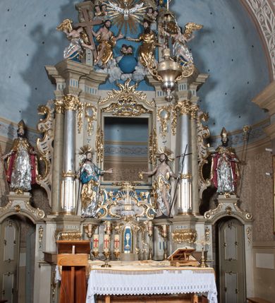 Zdjęcie nr 13: Nastawa ołtarzowa architektoniczna, jednoosiowa, jednokondygnacyjna ze zwieńczeniem, z bramkami i z rocaille'owymi uszami. Tabernakulum w formie zbliżonej do prostopadłościennej skrzynki, na wklęsło-wypukłym rzucie, ustawione na niskim cokole, o wypukłej ściance frontowej. Ujęte jest drewnianą obudową o wklęsłych bokach, flankowane parą ustawionych pod kątem pilastrów, których trzony przyjmują formę wolut. Zwieńczenie tabernakulum w formie niewielkiego szczytu, na którym ustawiona jest pasyjka. Całość ujęta po bokach rocaille'owymi wolutami. Drzwiczki w kształcie stojącego prostokąta, zamknięte łukiem nadwieszonym. W polu, na niebieskim tle malowane przedstawienie kielicha z hostią. Nastawa ustawiona na zdwojonym cokole, o  wklęsłym przebiegu, ujęta dwiema parami kolumn na tle zwielokrotnionych pilastrów. Podpory dźwigają pełne belkowanie, wyłamane pośrodku. Pomiędzy podporami figury: po lewej św. Józefa, a po prawej św. Jana Chrzciciela, nad bramkami: po lewej św. Wojciecha, a po prawej św. Stanisława. Nad tabernakulum rocaille'owy kartusz z łacińską inskrypcją w polu. W polu środkowym nisza w formie stojącego prostokąta, zamkniętego łukiem wklęsło-wypukłym z uskokami z obrazem „Święta Rozmowa”. Zwieńczenie w formie zbliżonej do stojącego prostokąta ujętego po bokach dwoma pilastrami, bogato ozdobione dekoracją rzeźbiarską. W polu przedstawienie Trójcy Świętej, flankowane przez parę aniołów siedzących na wolutowych przyczółkach. Całość zamyka para aniołków ze srebrną wstęgą z napisem „GLORIA IN EXCELSIS DEO”. Bramki z drzwiczkami w formie stojącego prostokąta, flankowane węgarami, zamknięte wolutowym łukiem z zatkniętymi na szczycie rzeźbami świętych. Drzwiczki dekorowane w polu dwiema płycinami. Całość nastawy silnie dekorowana ornamentyką rokokową. Kolumny ustawione na bazach, z kompozytowymi kapitelami, wewnętrzne srebrzone, do jednej trzeciej wysokości zdobione kanelowaniem zamkniętym pierścieniem, kolumny zewnętrzne marmoryzowane. Struktura polichromowana w kolorze beżowym, profilowania marmoryzowane, ornamentyka złocona.