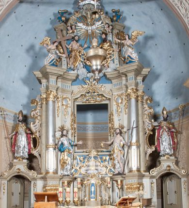 Zdjęcie nr 1: Nastawa ołtarzowa architektoniczna, jednoosiowa, jednokondygnacyjna ze zwieńczeniem, z bramkami i z rocaille&#039;owymi uszami. Tabernakulum w formie zbliżonej do prostopadłościennej skrzynki, na wklęsło-wypukłym rzucie, ustawione na niskim cokole, o wypukłej ściance frontowej. Ujęte jest drewnianą obudową o wklęsłych bokach, flankowane parą ustawionych pod kątem pilastrów, których trzony przyjmują formę wolut. Zwieńczenie tabernakulum w formie niewielkiego szczytu, na którym ustawiona jest pasyjka. Całość ujęta po bokach rocaille&#039;owymi wolutami. Drzwiczki w kształcie stojącego prostokąta, zamknięte łukiem nadwieszonym. W polu, na niebieskim tle malowane przedstawienie kielicha z hostią. Nastawa ustawiona na zdwojonym cokole, o  wklęsłym przebiegu, ujęta dwiema parami kolumn na tle zwielokrotnionych pilastrów. Podpory dźwigają pełne belkowanie, wyłamane pośrodku. Pomiędzy podporami figury: po lewej św. Józefa, a po prawej św. Jana Chrzciciela, nad bramkami: po lewej św. Wojciecha, a po prawej św. Stanisława. Nad tabernakulum rocaille&#039;owy kartusz z łacińską inskrypcją w polu. W polu środkowym nisza w formie stojącego prostokąta, zamkniętego łukiem wklęsło-wypukłym z uskokami z obrazem „Święta Rozmowa”. Zwieńczenie w formie zbliżonej do stojącego prostokąta ujętego po bokach dwoma pilastrami, bogato ozdobione dekoracją rzeźbiarską. W polu przedstawienie Trójcy Świętej, flankowane przez parę aniołów siedzących na wolutowych przyczółkach. Całość zamyka para aniołków ze srebrną wstęgą z napisem „GLORIA IN EXCELSIS DEO”. Bramki z drzwiczkami w formie stojącego prostokąta, flankowane węgarami, zamknięte wolutowym łukiem z zatkniętymi na szczycie rzeźbami świętych. Drzwiczki dekorowane w polu dwiema płycinami. Całość nastawy silnie dekorowana ornamentyką rokokową. Kolumny ustawione na bazach, z kompozytowymi kapitelami, wewnętrzne srebrzone, do jednej trzeciej wysokości zdobione kanelowaniem zamkniętym pierścieniem, kolumny zewnętrzne marmoryzowane. Struktura polichromowana w kolorze beżowym, profilowania marmoryzowane, ornamentyka złocona.