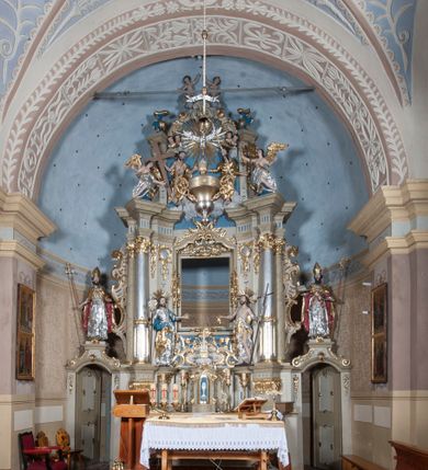 Zdjęcie nr 2: Nastawa ołtarzowa architektoniczna, jednoosiowa, jednokondygnacyjna ze zwieńczeniem, z bramkami i z rocaille'owymi uszami. Tabernakulum w formie zbliżonej do prostopadłościennej skrzynki, na wklęsło-wypukłym rzucie, ustawione na niskim cokole, o wypukłej ściance frontowej. Ujęte jest drewnianą obudową o wklęsłych bokach, flankowane parą ustawionych pod kątem pilastrów, których trzony przyjmują formę wolut. Zwieńczenie tabernakulum w formie niewielkiego szczytu, na którym ustawiona jest pasyjka. Całość ujęta po bokach rocaille'owymi wolutami. Drzwiczki w kształcie stojącego prostokąta, zamknięte łukiem nadwieszonym. W polu, na niebieskim tle malowane przedstawienie kielicha z hostią. Nastawa ustawiona na zdwojonym cokole, o  wklęsłym przebiegu, ujęta dwiema parami kolumn na tle zwielokrotnionych pilastrów. Podpory dźwigają pełne belkowanie, wyłamane pośrodku. Pomiędzy podporami figury: po lewej św. Józefa, a po prawej św. Jana Chrzciciela, nad bramkami: po lewej św. Wojciecha, a po prawej św. Stanisława. Nad tabernakulum rocaille'owy kartusz z łacińską inskrypcją w polu. W polu środkowym nisza w formie stojącego prostokąta, zamkniętego łukiem wklęsło-wypukłym z uskokami z obrazem „Święta Rozmowa”. Zwieńczenie w formie zbliżonej do stojącego prostokąta ujętego po bokach dwoma pilastrami, bogato ozdobione dekoracją rzeźbiarską. W polu przedstawienie Trójcy Świętej, flankowane przez parę aniołów siedzących na wolutowych przyczółkach. Całość zamyka para aniołków ze srebrną wstęgą z napisem „GLORIA IN EXCELSIS DEO”. Bramki z drzwiczkami w formie stojącego prostokąta, flankowane węgarami, zamknięte wolutowym łukiem z zatkniętymi na szczycie rzeźbami świętych. Drzwiczki dekorowane w polu dwiema płycinami. Całość nastawy silnie dekorowana ornamentyką rokokową. Kolumny ustawione na bazach, z kompozytowymi kapitelami, wewnętrzne srebrzone, do jednej trzeciej wysokości zdobione kanelowaniem zamkniętym pierścieniem, kolumny zewnętrzne marmoryzowane. Struktura polichromowana w kolorze beżowym, profilowania marmoryzowane, ornamentyka złocona.