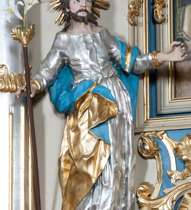 Zdjęcie nr 10: Nastawa ołtarzowa architektoniczna, jednoosiowa, jednokondygnacyjna ze zwieńczeniem, z bramkami i z rocaille'owymi uszami. Tabernakulum w formie zbliżonej do prostopadłościennej skrzynki, na wklęsło-wypukłym rzucie, ustawione na niskim cokole, o wypukłej ściance frontowej. Ujęte jest drewnianą obudową o wklęsłych bokach, flankowane parą ustawionych pod kątem pilastrów, których trzony przyjmują formę wolut. Zwieńczenie tabernakulum w formie niewielkiego szczytu, na którym ustawiona jest pasyjka. Całość ujęta po bokach rocaille'owymi wolutami. Drzwiczki w kształcie stojącego prostokąta, zamknięte łukiem nadwieszonym. W polu, na niebieskim tle malowane przedstawienie kielicha z hostią. Nastawa ustawiona na zdwojonym cokole, o  wklęsłym przebiegu, ujęta dwiema parami kolumn na tle zwielokrotnionych pilastrów. Podpory dźwigają pełne belkowanie, wyłamane pośrodku. Pomiędzy podporami figury: po lewej św. Józefa, a po prawej św. Jana Chrzciciela, nad bramkami: po lewej św. Wojciecha, a po prawej św. Stanisława. Nad tabernakulum rocaille'owy kartusz z łacińską inskrypcją w polu. W polu środkowym nisza w formie stojącego prostokąta, zamkniętego łukiem wklęsło-wypukłym z uskokami z obrazem „Święta Rozmowa”. Zwieńczenie w formie zbliżonej do stojącego prostokąta ujętego po bokach dwoma pilastrami, bogato ozdobione dekoracją rzeźbiarską. W polu przedstawienie Trójcy Świętej, flankowane przez parę aniołów siedzących na wolutowych przyczółkach. Całość zamyka para aniołków ze srebrną wstęgą z napisem „GLORIA IN EXCELSIS DEO”. Bramki z drzwiczkami w formie stojącego prostokąta, flankowane węgarami, zamknięte wolutowym łukiem z zatkniętymi na szczycie rzeźbami świętych. Drzwiczki dekorowane w polu dwiema płycinami. Całość nastawy silnie dekorowana ornamentyką rokokową. Kolumny ustawione na bazach, z kompozytowymi kapitelami, wewnętrzne srebrzone, do jednej trzeciej wysokości zdobione kanelowaniem zamkniętym pierścieniem, kolumny zewnętrzne marmoryzowane. Struktura polichromowana w kolorze beżowym, profilowania marmoryzowane, ornamentyka złocona.