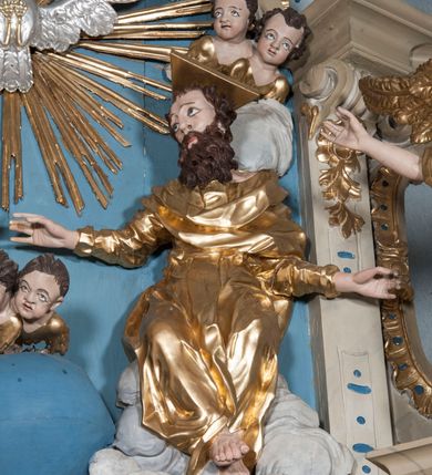 Zdjęcie nr 10: Zwieńczenie ołtarza w formie rzeźbiarskiej grupy z przedstawieniem Trójcy Świętej na tle obłoków w dolnej części kompozycji i promienistej glorii z gołębicą Ducha Świętego w górnej części. Po prawej stronie figura Boga Ojca, zwróconego delikatnie w swoją prawą stronę, w pozycji siedzącej, ze skrzyżowanymi nogami, z rękami rozłożonymi szeroko na boki. Twarz okolona długą i bujną, ciemnobrązową brodą, nos długi i szeroki, oczy duże skierowane na Chrystusa, nad głową trójkątny nimb. Bóg Ojciec jest ubrany w długą, złoconą suknię z długimi rękawami. Po lewej stronie figura Chrystusa zwróconego delikatnie w lewo, w pozycji siedzącej z rękami ugiętymi w łokciach i rozłożonymi na boki, w prawej ręce trzyma duży krzyż. Twarz szeroka, usta rozchylone, nos długi i szeroki, oczy skierowane na Boga Ojca, głowa okolona krótką brodą. Wokół głowy zachowana jedna wiązka promieni z nimbu krzyżowego. Nad nimi gołębica Ducha Świętego z rozpostartymi szeroko skrzydłami na tle promienistej glorii złożonej z wiązek promieni o nierównej długości. Poniżej błękitny glob ziemski, a nad nim oraz w górnej części kompozycji pary uskrzydlonych główek anielskich. Po bokach zwieńczenia dwie figury aniołów usadzone na wolutowych przyczółkach. Anioły ukazane w pozycji klęczącej o silnie wygiętej sylwetce z rękami rozłożonymi szeroko na boki i uniesionymi ku górze. Ubrane w srebrzone szaty zawieszone na wąskim pasku, odsłaniające ramiona i nogi, u pleców duże złocone skrzydła. Całość zwieńczona figurami dwóch małych aniołków trzymających wstęgę z napisem „GLORIA IN EXCELSIS DEO”. Polichromia w odsłoniętych partiach ciała naturalistyczna, szaty i detale złocone i srebrzone.
