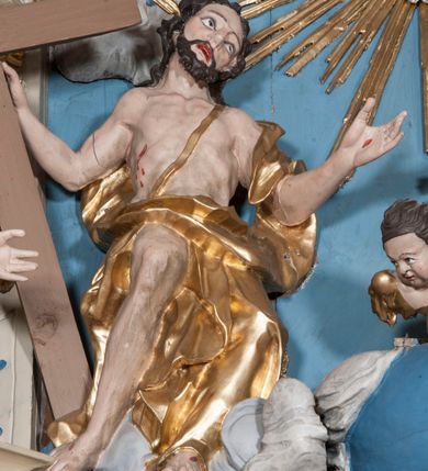 Zdjęcie nr 9: Zwieńczenie ołtarza w formie rzeźbiarskiej grupy z przedstawieniem Trójcy Świętej na tle obłoków w dolnej części kompozycji i promienistej glorii z gołębicą Ducha Świętego w górnej części. Po prawej stronie figura Boga Ojca, zwróconego delikatnie w swoją prawą stronę, w pozycji siedzącej, ze skrzyżowanymi nogami, z rękami rozłożonymi szeroko na boki. Twarz okolona długą i bujną, ciemnobrązową brodą, nos długi i szeroki, oczy duże skierowane na Chrystusa, nad głową trójkątny nimb. Bóg Ojciec jest ubrany w długą, złoconą suknię z długimi rękawami. Po lewej stronie figura Chrystusa zwróconego delikatnie w lewo, w pozycji siedzącej z rękami ugiętymi w łokciach i rozłożonymi na boki, w prawej ręce trzyma duży krzyż. Twarz szeroka, usta rozchylone, nos długi i szeroki, oczy skierowane na Boga Ojca, głowa okolona krótką brodą. Wokół głowy zachowana jedna wiązka promieni z nimbu krzyżowego. Nad nimi gołębica Ducha Świętego z rozpostartymi szeroko skrzydłami na tle promienistej glorii złożonej z wiązek promieni o nierównej długości. Poniżej błękitny glob ziemski, a nad nim oraz w górnej części kompozycji pary uskrzydlonych główek anielskich. Po bokach zwieńczenia dwie figury aniołów usadzone na wolutowych przyczółkach. Anioły ukazane w pozycji klęczącej o silnie wygiętej sylwetce z rękami rozłożonymi szeroko na boki i uniesionymi ku górze. Ubrane w srebrzone szaty zawieszone na wąskim pasku, odsłaniające ramiona i nogi, u pleców duże złocone skrzydła. Całość zwieńczona figurami dwóch małych aniołków trzymających wstęgę z napisem „GLORIA IN EXCELSIS DEO”. Polichromia w odsłoniętych partiach ciała naturalistyczna, szaty i detale złocone i srebrzone.
