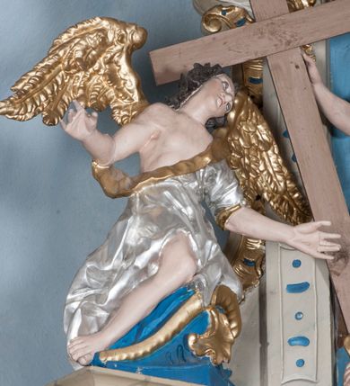 Zdjęcie nr 12: Zwieńczenie ołtarza w formie rzeźbiarskiej grupy z przedstawieniem Trójcy Świętej na tle obłoków w dolnej części kompozycji i promienistej glorii z gołębicą Ducha Świętego w górnej części. Po prawej stronie figura Boga Ojca, zwróconego delikatnie w swoją prawą stronę, w pozycji siedzącej, ze skrzyżowanymi nogami, z rękami rozłożonymi szeroko na boki. Twarz okolona długą i bujną, ciemnobrązową brodą, nos długi i szeroki, oczy duże skierowane na Chrystusa, nad głową trójkątny nimb. Bóg Ojciec jest ubrany w długą, złoconą suknię z długimi rękawami. Po lewej stronie figura Chrystusa zwróconego delikatnie w lewo, w pozycji siedzącej z rękami ugiętymi w łokciach i rozłożonymi na boki, w prawej ręce trzyma duży krzyż. Twarz szeroka, usta rozchylone, nos długi i szeroki, oczy skierowane na Boga Ojca, głowa okolona krótką brodą. Wokół głowy zachowana jedna wiązka promieni z nimbu krzyżowego. Nad nimi gołębica Ducha Świętego z rozpostartymi szeroko skrzydłami na tle promienistej glorii złożonej z wiązek promieni o nierównej długości. Poniżej błękitny glob ziemski, a nad nim oraz w górnej części kompozycji pary uskrzydlonych główek anielskich. Po bokach zwieńczenia dwie figury aniołów usadzone na wolutowych przyczółkach. Anioły ukazane w pozycji klęczącej o silnie wygiętej sylwetce z rękami rozłożonymi szeroko na boki i uniesionymi ku górze. Ubrane w srebrzone szaty zawieszone na wąskim pasku, odsłaniające ramiona i nogi, u pleców duże złocone skrzydła. Całość zwieńczona figurami dwóch małych aniołków trzymających wstęgę z napisem „GLORIA IN EXCELSIS DEO”. Polichromia w odsłoniętych partiach ciała naturalistyczna, szaty i detale złocone i srebrzone.
