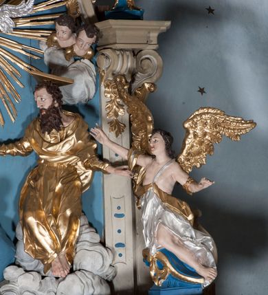 Zdjęcie nr 8: Zwieńczenie ołtarza w formie rzeźbiarskiej grupy z przedstawieniem Trójcy Świętej na tle obłoków w dolnej części kompozycji i promienistej glorii z gołębicą Ducha Świętego w górnej części. Po prawej stronie figura Boga Ojca, zwróconego delikatnie w swoją prawą stronę, w pozycji siedzącej, ze skrzyżowanymi nogami, z rękami rozłożonymi szeroko na boki. Twarz okolona długą i bujną, ciemnobrązową brodą, nos długi i szeroki, oczy duże skierowane na Chrystusa, nad głową trójkątny nimb. Bóg Ojciec jest ubrany w długą, złoconą suknię z długimi rękawami. Po lewej stronie figura Chrystusa zwróconego delikatnie w lewo, w pozycji siedzącej z rękami ugiętymi w łokciach i rozłożonymi na boki, w prawej ręce trzyma duży krzyż. Twarz szeroka, usta rozchylone, nos długi i szeroki, oczy skierowane na Boga Ojca, głowa okolona krótką brodą. Wokół głowy zachowana jedna wiązka promieni z nimbu krzyżowego. Nad nimi gołębica Ducha Świętego z rozpostartymi szeroko skrzydłami na tle promienistej glorii złożonej z wiązek promieni o nierównej długości. Poniżej błękitny glob ziemski, a nad nim oraz w górnej części kompozycji pary uskrzydlonych główek anielskich. Po bokach zwieńczenia dwie figury aniołów usadzone na wolutowych przyczółkach. Anioły ukazane w pozycji klęczącej o silnie wygiętej sylwetce z rękami rozłożonymi szeroko na boki i uniesionymi ku górze. Ubrane w srebrzone szaty zawieszone na wąskim pasku, odsłaniające ramiona i nogi, u pleców duże złocone skrzydła. Całość zwieńczona figurami dwóch małych aniołków trzymających wstęgę z napisem „GLORIA IN EXCELSIS DEO”. Polichromia w odsłoniętych partiach ciała naturalistyczna, szaty i detale złocone i srebrzone.
