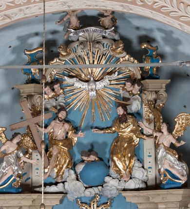 Zdjęcie nr 2: Zwieńczenie ołtarza w formie rzeźbiarskiej grupy z przedstawieniem Trójcy Świętej na tle obłoków w dolnej części kompozycji i promienistej glorii z gołębicą Ducha Świętego w górnej części. Po prawej stronie figura Boga Ojca, zwróconego delikatnie w swoją prawą stronę, w pozycji siedzącej, ze skrzyżowanymi nogami, z rękami rozłożonymi szeroko na boki. Twarz okolona długą i bujną, ciemnobrązową brodą, nos długi i szeroki, oczy duże skierowane na Chrystusa, nad głową trójkątny nimb. Bóg Ojciec jest ubrany w długą, złoconą suknię z długimi rękawami. Po lewej stronie figura Chrystusa zwróconego delikatnie w lewo, w pozycji siedzącej z rękami ugiętymi w łokciach i rozłożonymi na boki, w prawej ręce trzyma duży krzyż. Twarz szeroka, usta rozchylone, nos długi i szeroki, oczy skierowane na Boga Ojca, głowa okolona krótką brodą. Wokół głowy zachowana jedna wiązka promieni z nimbu krzyżowego. Nad nimi gołębica Ducha Świętego z rozpostartymi szeroko skrzydłami na tle promienistej glorii złożonej z wiązek promieni o nierównej długości. Poniżej błękitny glob ziemski, a nad nim oraz w górnej części kompozycji pary uskrzydlonych główek anielskich. Po bokach zwieńczenia dwie figury aniołów usadzone na wolutowych przyczółkach. Anioły ukazane w pozycji klęczącej o silnie wygiętej sylwetce z rękami rozłożonymi szeroko na boki i uniesionymi ku górze. Ubrane w srebrzone szaty zawieszone na wąskim pasku, odsłaniające ramiona i nogi, u pleców duże złocone skrzydła. Całość zwieńczona figurami dwóch małych aniołków trzymających wstęgę z napisem „GLORIA IN EXCELSIS DEO”. Polichromia w odsłoniętych partiach ciała naturalistyczna, szaty i detale złocone i srebrzone.
