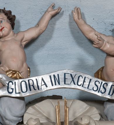 Zdjęcie nr 4: Zwieńczenie ołtarza w formie rzeźbiarskiej grupy z przedstawieniem Trójcy Świętej na tle obłoków w dolnej części kompozycji i promienistej glorii z gołębicą Ducha Świętego w górnej części. Po prawej stronie figura Boga Ojca, zwróconego delikatnie w swoją prawą stronę, w pozycji siedzącej, ze skrzyżowanymi nogami, z rękami rozłożonymi szeroko na boki. Twarz okolona długą i bujną, ciemnobrązową brodą, nos długi i szeroki, oczy duże skierowane na Chrystusa, nad głową trójkątny nimb. Bóg Ojciec jest ubrany w długą, złoconą suknię z długimi rękawami. Po lewej stronie figura Chrystusa zwróconego delikatnie w lewo, w pozycji siedzącej z rękami ugiętymi w łokciach i rozłożonymi na boki, w prawej ręce trzyma duży krzyż. Twarz szeroka, usta rozchylone, nos długi i szeroki, oczy skierowane na Boga Ojca, głowa okolona krótką brodą. Wokół głowy zachowana jedna wiązka promieni z nimbu krzyżowego. Nad nimi gołębica Ducha Świętego z rozpostartymi szeroko skrzydłami na tle promienistej glorii złożonej z wiązek promieni o nierównej długości. Poniżej błękitny glob ziemski, a nad nim oraz w górnej części kompozycji pary uskrzydlonych główek anielskich. Po bokach zwieńczenia dwie figury aniołów usadzone na wolutowych przyczółkach. Anioły ukazane w pozycji klęczącej o silnie wygiętej sylwetce z rękami rozłożonymi szeroko na boki i uniesionymi ku górze. Ubrane w srebrzone szaty zawieszone na wąskim pasku, odsłaniające ramiona i nogi, u pleców duże złocone skrzydła. Całość zwieńczona figurami dwóch małych aniołków trzymających wstęgę z napisem „GLORIA IN EXCELSIS DEO”. Polichromia w odsłoniętych partiach ciała naturalistyczna, szaty i detale złocone i srebrzone.
