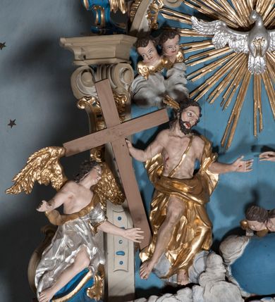 Zdjęcie nr 7: Zwieńczenie ołtarza w formie rzeźbiarskiej grupy z przedstawieniem Trójcy Świętej na tle obłoków w dolnej części kompozycji i promienistej glorii z gołębicą Ducha Świętego w górnej części. Po prawej stronie figura Boga Ojca, zwróconego delikatnie w swoją prawą stronę, w pozycji siedzącej, ze skrzyżowanymi nogami, z rękami rozłożonymi szeroko na boki. Twarz okolona długą i bujną, ciemnobrązową brodą, nos długi i szeroki, oczy duże skierowane na Chrystusa, nad głową trójkątny nimb. Bóg Ojciec jest ubrany w długą, złoconą suknię z długimi rękawami. Po lewej stronie figura Chrystusa zwróconego delikatnie w lewo, w pozycji siedzącej z rękami ugiętymi w łokciach i rozłożonymi na boki, w prawej ręce trzyma duży krzyż. Twarz szeroka, usta rozchylone, nos długi i szeroki, oczy skierowane na Boga Ojca, głowa okolona krótką brodą. Wokół głowy zachowana jedna wiązka promieni z nimbu krzyżowego. Nad nimi gołębica Ducha Świętego z rozpostartymi szeroko skrzydłami na tle promienistej glorii złożonej z wiązek promieni o nierównej długości. Poniżej błękitny glob ziemski, a nad nim oraz w górnej części kompozycji pary uskrzydlonych główek anielskich. Po bokach zwieńczenia dwie figury aniołów usadzone na wolutowych przyczółkach. Anioły ukazane w pozycji klęczącej o silnie wygiętej sylwetce z rękami rozłożonymi szeroko na boki i uniesionymi ku górze. Ubrane w srebrzone szaty zawieszone na wąskim pasku, odsłaniające ramiona i nogi, u pleców duże złocone skrzydła. Całość zwieńczona figurami dwóch małych aniołków trzymających wstęgę z napisem „GLORIA IN EXCELSIS DEO”. Polichromia w odsłoniętych partiach ciała naturalistyczna, szaty i detale złocone i srebrzone.
