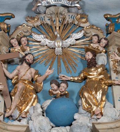 Zdjęcie nr 1: Zwieńczenie ołtarza w formie rzeźbiarskiej grupy z przedstawieniem Trójcy Świętej na tle obłoków w dolnej części kompozycji i promienistej glorii z gołębicą Ducha Świętego w górnej części. Po prawej stronie figura Boga Ojca, zwróconego delikatnie w swoją prawą stronę, w pozycji siedzącej, ze skrzyżowanymi nogami, z rękami rozłożonymi szeroko na boki. Twarz okolona długą i bujną, ciemnobrązową brodą, nos długi i szeroki, oczy duże skierowane na Chrystusa, nad głową trójkątny nimb. Bóg Ojciec jest ubrany w długą, złoconą suknię z długimi rękawami. Po lewej stronie figura Chrystusa zwróconego delikatnie w lewo, w pozycji siedzącej z rękami ugiętymi w łokciach i rozłożonymi na boki, w prawej ręce trzyma duży krzyż. Twarz szeroka, usta rozchylone, nos długi i szeroki, oczy skierowane na Boga Ojca, głowa okolona krótką brodą. Wokół głowy zachowana jedna wiązka promieni z nimbu krzyżowego. Nad nimi gołębica Ducha Świętego z rozpostartymi szeroko skrzydłami na tle promienistej glorii złożonej z wiązek promieni o nierównej długości. Poniżej błękitny glob ziemski, a nad nim oraz w górnej części kompozycji pary uskrzydlonych główek anielskich. Po bokach zwieńczenia dwie figury aniołów usadzone na wolutowych przyczółkach. Anioły ukazane w pozycji klęczącej o silnie wygiętej sylwetce z rękami rozłożonymi szeroko na boki i uniesionymi ku górze. Ubrane w srebrzone szaty zawieszone na wąskim pasku, odsłaniające ramiona i nogi, u pleców duże złocone skrzydła. Całość zwieńczona figurami dwóch małych aniołków trzymających wstęgę z napisem „GLORIA IN EXCELSIS DEO”. Polichromia w odsłoniętych partiach ciała naturalistyczna, szaty i detale złocone i srebrzone.
