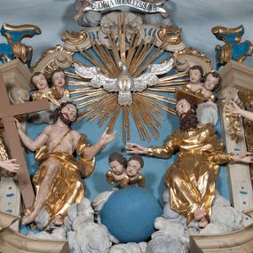 Zdjęcie nr 1: Zwieńczenie ołtarza w formie rzeźbiarskiej grupy z przedstawieniem Trójcy Świętej na tle obłoków w dolnej części kompozycji i promienistej glorii z gołębicą Ducha Świętego w górnej części. Po prawej stronie figura Boga Ojca, zwróconego delikatnie w swoją prawą stronę, w pozycji siedzącej, ze skrzyżowanymi nogami, z rękami rozłożonymi szeroko na boki. Twarz okolona długą i bujną, ciemnobrązową brodą, nos długi i szeroki, oczy duże skierowane na Chrystusa, nad głową trójkątny nimb. Bóg Ojciec jest ubrany w długą, złoconą suknię z długimi rękawami. Po lewej stronie figura Chrystusa zwróconego delikatnie w lewo, w pozycji siedzącej z rękami ugiętymi w łokciach i rozłożonymi na boki, w prawej ręce trzyma duży krzyż. Twarz szeroka, usta rozchylone, nos długi i szeroki, oczy skierowane na Boga Ojca, głowa okolona krótką brodą. Wokół głowy zachowana jedna wiązka promieni z nimbu krzyżowego. Nad nimi gołębica Ducha Świętego z rozpostartymi szeroko skrzydłami na tle promienistej glorii złożonej z wiązek promieni o nierównej długości. Poniżej błękitny glob ziemski, a nad nim oraz w górnej części kompozycji pary uskrzydlonych główek anielskich. Po bokach zwieńczenia dwie figury aniołów usadzone na wolutowych przyczółkach. Anioły ukazane w pozycji klęczącej o silnie wygiętej sylwetce z rękami rozłożonymi szeroko na boki i uniesionymi ku górze. Ubrane w srebrzone szaty zawieszone na wąskim pasku, odsłaniające ramiona i nogi, u pleców duże złocone skrzydła. Całość zwieńczona figurami dwóch małych aniołków trzymających wstęgę z napisem „GLORIA IN EXCELSIS DEO”. Polichromia w odsłoniętych partiach ciała naturalistyczna, szaty i detale złocone i srebrzone.
