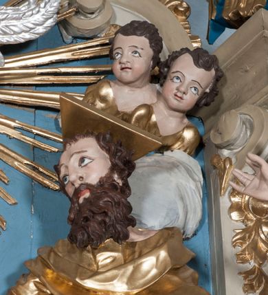 Zdjęcie nr 3: Zwieńczenie ołtarza w formie rzeźbiarskiej grupy z przedstawieniem Trójcy Świętej na tle obłoków w dolnej części kompozycji i promienistej glorii z gołębicą Ducha Świętego w górnej części. Po prawej stronie figura Boga Ojca, zwróconego delikatnie w swoją prawą stronę, w pozycji siedzącej, ze skrzyżowanymi nogami, z rękami rozłożonymi szeroko na boki. Twarz okolona długą i bujną, ciemnobrązową brodą, nos długi i szeroki, oczy duże skierowane na Chrystusa, nad głową trójkątny nimb. Bóg Ojciec jest ubrany w długą, złoconą suknię z długimi rękawami. Po lewej stronie figura Chrystusa zwróconego delikatnie w lewo, w pozycji siedzącej z rękami ugiętymi w łokciach i rozłożonymi na boki, w prawej ręce trzyma duży krzyż. Twarz szeroka, usta rozchylone, nos długi i szeroki, oczy skierowane na Boga Ojca, głowa okolona krótką brodą. Wokół głowy zachowana jedna wiązka promieni z nimbu krzyżowego. Nad nimi gołębica Ducha Świętego z rozpostartymi szeroko skrzydłami na tle promienistej glorii złożonej z wiązek promieni o nierównej długości. Poniżej błękitny glob ziemski, a nad nim oraz w górnej części kompozycji pary uskrzydlonych główek anielskich. Po bokach zwieńczenia dwie figury aniołów usadzone na wolutowych przyczółkach. Anioły ukazane w pozycji klęczącej o silnie wygiętej sylwetce z rękami rozłożonymi szeroko na boki i uniesionymi ku górze. Ubrane w srebrzone szaty zawieszone na wąskim pasku, odsłaniające ramiona i nogi, u pleców duże złocone skrzydła. Całość zwieńczona figurami dwóch małych aniołków trzymających wstęgę z napisem „GLORIA IN EXCELSIS DEO”. Polichromia w odsłoniętych partiach ciała naturalistyczna, szaty i detale złocone i srebrzone.
