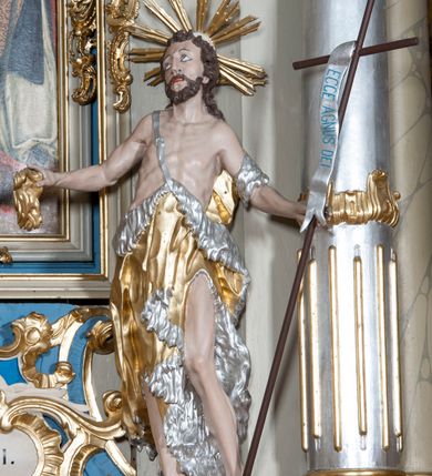 Zdjęcie nr 2: Rzeźba ustawiona na niskim postumencie. Święty ukazany w postawie stojącej w kontrapoście, z lewą nogą ugiętą w kolanie, z krzyżem w lewej dłoni oraz niezidentyfikowanym przedmiotem w prawej dłoni (muszla?), z głową zwróconą delikatnie w prawo. Twarz szczupła, okolona krótką, ciemnobrązową brodą, długi i wąski nos, wzrok skierowany w prawo i ku górze. Włosy długie, ciemnobrązowe, kręcone, spływające na plecy. Wokół głowy promienisty nimb. Święty jest ubrany w skórę wielbłądzią zawieszoną na pasku na prawym ramieniu, odsłaniającą nagi tors i lewą nogę. Krzyż prosty z pionową banderolą z napisem „ECCE AGNUS DEI”. Polichromia w odsłoniętych partiach ciała naturalistyczna, szaty i detale złocone i srebrzone.
