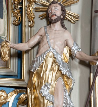 Zdjęcie nr 4: Rzeźba ustawiona na niskim postumencie. Święty ukazany w postawie stojącej w kontrapoście, z lewą nogą ugiętą w kolanie, z krzyżem w lewej dłoni oraz niezidentyfikowanym przedmiotem w prawej dłoni (muszla?), z głową zwróconą delikatnie w prawo. Twarz szczupła, okolona krótką, ciemnobrązową brodą, długi i wąski nos, wzrok skierowany w prawo i ku górze. Włosy długie, ciemnobrązowe, kręcone, spływające na plecy. Wokół głowy promienisty nimb. Święty jest ubrany w skórę wielbłądzią zawieszoną na pasku na prawym ramieniu, odsłaniającą nagi tors i lewą nogę. Krzyż prosty z pionową banderolą z napisem „ECCE AGNUS DEI”. Polichromia w odsłoniętych partiach ciała naturalistyczna, szaty i detale złocone i srebrzone.
