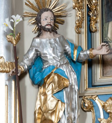 Zdjęcie nr 1: Rzeźba przedstawiająca św. Józefa na niskim cokole. Święty ukazany w pozycji stojącej o esowato wygiętej sylwetce, z gałązką białej lilii w prawej dłoni, z lewą uniesioną na wysokości piersi i odsuniętą w bok. Twarz szeroka, okolona krótką i kręconą brodą, zakończoną dwoma spiralnie skręconymi puklami, nos długi i szeroki, oczy głęboko osadzone, skierowane ku górze, okolone zmarszczonymi brwiami, usta delikatnie rozchylone, włosy długie i ciemnobrązowe opadające na plecy i ramiona. Wokół głowy promienisty nimb złożony z wiązek promieni nierównej długości. Święty ubrany w długą, srebrzoną suknię z długimi rękawami, odsłaniającą dekolt oraz złocony płaszcz z niebieską podszewką, na stopach sandały. Polichromia w odsłoniętych partiach ciała naturalistyczna. 

