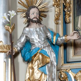 Zdjęcie nr 1: Rzeźba przedstawiająca św. Józefa na niskim cokole. Święty ukazany w pozycji stojącej o esowato wygiętej sylwetce, z gałązką białej lilii w prawej dłoni, z lewą uniesioną na wysokości piersi i odsuniętą w bok. Twarz szeroka, okolona krótką i kręconą brodą, zakończoną dwoma spiralnie skręconymi puklami, nos długi i szeroki, oczy głęboko osadzone, skierowane ku górze, okolone zmarszczonymi brwiami, usta delikatnie rozchylone, włosy długie i ciemnobrązowe opadające na plecy i ramiona. Wokół głowy promienisty nimb złożony z wiązek promieni nierównej długości. Święty ubrany w długą, srebrzoną suknię z długimi rękawami, odsłaniającą dekolt oraz złocony płaszcz z niebieską podszewką, na stopach sandały. Polichromia w odsłoniętych partiach ciała naturalistyczna. 
