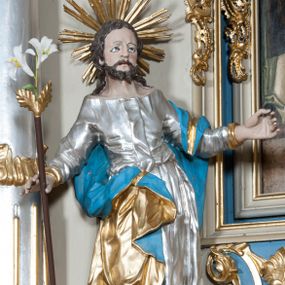 Zdjęcie nr 1: Rzeźba przedstawiająca św. Józefa na niskim cokole. Święty ukazany w pozycji stojącej o esowato wygiętej sylwetce, z gałązką białej lilii w prawej dłoni, z lewą uniesioną na wysokości piersi i odsuniętą w bok. Twarz szeroka, okolona krótką i kręconą brodą, zakończoną dwoma spiralnie skręconymi puklami, nos długi i szeroki, oczy głęboko osadzone, skierowane ku górze, okolone zmarszczonymi brwiami, usta delikatnie rozchylone, włosy długie i ciemnobrązowe opadające na plecy i ramiona. Wokół głowy promienisty nimb złożony z wiązek promieni nierównej długości. Święty ubrany w długą, srebrzoną suknię z długimi rękawami, odsłaniającą dekolt oraz złocony płaszcz z niebieską podszewką, na stopach sandały. Polichromia w odsłoniętych partiach ciała naturalistyczna. 
