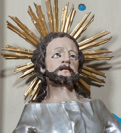 Zdjęcie nr 2: Rzeźba przedstawiająca św. Józefa na niskim cokole. Święty ukazany w pozycji stojącej o esowato wygiętej sylwetce, z gałązką białej lilii w prawej dłoni, z lewą uniesioną na wysokości piersi i odsuniętą w bok. Twarz szeroka, okolona krótką i kręconą brodą, zakończoną dwoma spiralnie skręconymi puklami, nos długi i szeroki, oczy głęboko osadzone, skierowane ku górze, okolone zmarszczonymi brwiami, usta delikatnie rozchylone, włosy długie i ciemnobrązowe opadające na plecy i ramiona. Wokół głowy promienisty nimb złożony z wiązek promieni nierównej długości. Święty ubrany w długą, srebrzoną suknię z długimi rękawami, odsłaniającą dekolt oraz złocony płaszcz z niebieską podszewką, na stopach sandały. Polichromia w odsłoniętych partiach ciała naturalistyczna. 
