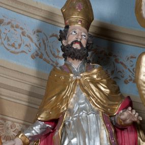 Zdjęcie nr 1: Rzeźba ustawiona na niskim, prostopadłościennym cokole przedstawiająca św. Stanisława w postawie stojącej, w delikatnym kontrapoście, z prawa nogą ugiętą w kolanie, z krzyżem w prawej dłoni, z lewą ręką odsuniętą delikatnie w bok, z głową skierowaną ku górze. Twarz podłużna, okolona średniej długości, ciemnobrązową, bujną brodą, oczy skierowane ku górze, nos długi i szeroki, włosy średniej długości, kręcone. Święty jest ubrany w albę z długimi rękawami oraz złoconą kapę z czerwoną podszewką spiętą na piersi klamrą, spod kapy widoczna czerwona stuła, na stopach złocone buty, a na głowie infuła. Polichromia w odsłoniętych partiach ciała naturalistyczna. 