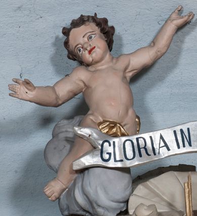 Zdjęcie nr 1: Rzeźby aniołów usytuowano na szczycie ołtarza głównego. Aniołki są ukazane w odbiciu lustrzanym, usadzone na szarych obłokach o silnie wygiętej sylwetce z rękami wzniesionymi ku górze, z przechylonymi na bok głowami. Mają twarze szerokie o pulchnych policzkach z krótkimi i szerokimi noskami, wąskimi ustami, z wyraźnie zaznaczonymi podbródkami i dużymi oczami skierowanymi ku górze. Włosy krótkie, ciemnobrązowe, kędzierzawe, zasłaniające uszy. Sylwetki o obfitych kształtach. Aniołki są przesłonięte w biodrach złotą, wąską, drapowaną tkaniną. Pomiędzy aniołkami rozpięta banderola z napisem „GLORIA IN EXCELSIS DEO”. Polichromia w odsłoniętych partiach ciała naturalistyczna, tkaniny złocone, banderola srebrzona.