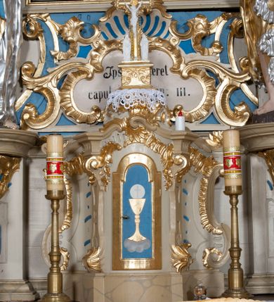 Zdjęcie nr 1: Tabernakulum w formie zbliżonej do prostopadłościennej skrzynki, na wklęsło-wypukłym rzucie, ustawione na niskim cokole, o wypukłej ściance frontowej, ujęte drewnianą obudową o wklęsłych bokach, flankowane parą ustawionych pod kątem pilastrów, których trzony przyjmują formę wolut. Zwieńczenie tabernakulum w formie niewielkiego szczytu, na którym ustawiona jest pasyjka. Całość ujęta po bokach rocaille'owymi wolutami. Drzwiczki w kształcie stojącego prostokąta, zamknięte łukiem nadwieszonym. W polu, na niebieskim tle malowane przedstawienie kielicha z hostią. Struktura polichromowana na kolor jasnobeżowy, detale na kolor ciemnobeżowy, cokół marmoryzowany, ornamentyka złocona.