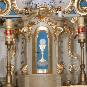 Zdjęcie nr 1: Tabernakulum w formie zbliżonej do prostopadłościennej skrzynki, na wklęsło-wypukłym rzucie, ustawione na niskim cokole, o wypukłej ściance frontowej, ujęte drewnianą obudową o wklęsłych bokach, flankowane parą ustawionych pod kątem pilastrów, których trzony przyjmują formę wolut. Zwieńczenie tabernakulum w formie niewielkiego szczytu, na którym ustawiona jest pasyjka. Całość ujęta po bokach rocaille'owymi wolutami. Drzwiczki w kształcie stojącego prostokąta, zamknięte łukiem nadwieszonym. W polu, na niebieskim tle malowane przedstawienie kielicha z hostią. Struktura polichromowana na kolor jasnobeżowy, detale na kolor ciemnobeżowy, cokół marmoryzowany, ornamentyka złocona.