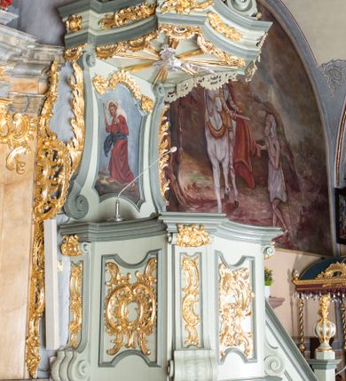 Zdjęcie nr 1: Ambona przyścienna, wisząca, z dostępem od strony prezbiterium w postaci jednobiegowych schodów z pełną balustradą dekorowaną dwoma płycinami w kształcie rombów oraz poręczą w formie gzymsu, zakończoną filarem z rokokowym wazonem. Kosz na planie kwadratu o ściętych narożach i wklęsłych bokach, na profilowanym cokole; u spodu podwieszony ostrosłup o wklęsłych bokach, z wolutami w narożach, zakończony szyszką w formie rocaille'u. Ścianki kosza flankowane pilastrami w formie spływów wolutowych z płyciną pośrodku w kształcie stojącego prostokąta o wklęsłych górnych i spodnich bokach; w polu ścianek kosza płyciny analogiczne jak na spływach wolutowych z dekoracją rocaille'ową w polu. Parapet w postaci profilowanego gzymsu. Zaplecek w kształcie stojącego prostokąta o wklęsłych bokach, ujętych spływami wolutowymi. W polu obraz Chrystusa Dobrego Pasterza umieszczony w płycinie w formie stojącego prostokąta o wklęsłych bokach, zamkniętego łukiem nadwieszonym z uskokami; u góry rocaille i rozety. Zaplecek flankowany uszami w postaci strzępionych liści. Baldachim na planie analogicznym jak kosz w postaci pełnego belkowania dekorowanego ornamentem rocaille'owym. Belkowanie stanowi podstawę dla czterech wolutowych kabłąków, dźwigających cokół z rokokowym wazonem. U spodu baldachimu rzeźbiona gołębica Ducha Świętego w promienistej glorii. Struktura polichromowana w kolorze seledynowym z ciemniejszymi profilami, ornamentyka złocona, gołębica srebrzona.