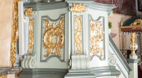 Zdjęcie nr 1: Ambona przyścienna, wisząca, z dostępem od strony prezbiterium w postaci jednobiegowych schodów z pełną balustradą dekorowaną dwoma płycinami w kształcie rombów oraz poręczą w formie gzymsu, zakończoną filarem z rokokowym wazonem. Kosz na planie kwadratu o ściętych narożach i wklęsłych bokach, na profilowanym cokole; u spodu podwieszony ostrosłup o wklęsłych bokach, z wolutami w narożach, zakończony szyszką w formie rocaille&#039;u. Ścianki kosza flankowane pilastrami w formie spływów wolutowych z płyciną pośrodku w kształcie stojącego prostokąta o wklęsłych górnych i spodnich bokach; w polu ścianek kosza płyciny analogiczne jak na spływach wolutowych z dekoracją rocaille&#039;ową w polu. Parapet w postaci profilowanego gzymsu. Zaplecek w kształcie stojącego prostokąta o wklęsłych bokach, ujętych spływami wolutowymi. W polu obraz Chrystusa Dobrego Pasterza umieszczony w płycinie w formie stojącego prostokąta o wklęsłych bokach, zamkniętego łukiem nadwieszonym z uskokami; u góry rocaille i rozety. Zaplecek flankowany uszami w postaci strzępionych liści. Baldachim na planie analogicznym jak kosz w postaci pełnego belkowania dekorowanego ornamentem rocaille&#039;owym. Belkowanie stanowi podstawę dla czterech wolutowych kabłąków, dźwigających cokół z rokokowym wazonem. U spodu baldachimu rzeźbiona gołębica Ducha Świętego w promienistej glorii. Struktura polichromowana w kolorze seledynowym z ciemniejszymi profilami, ornamentyka złocona, gołębica srebrzona.